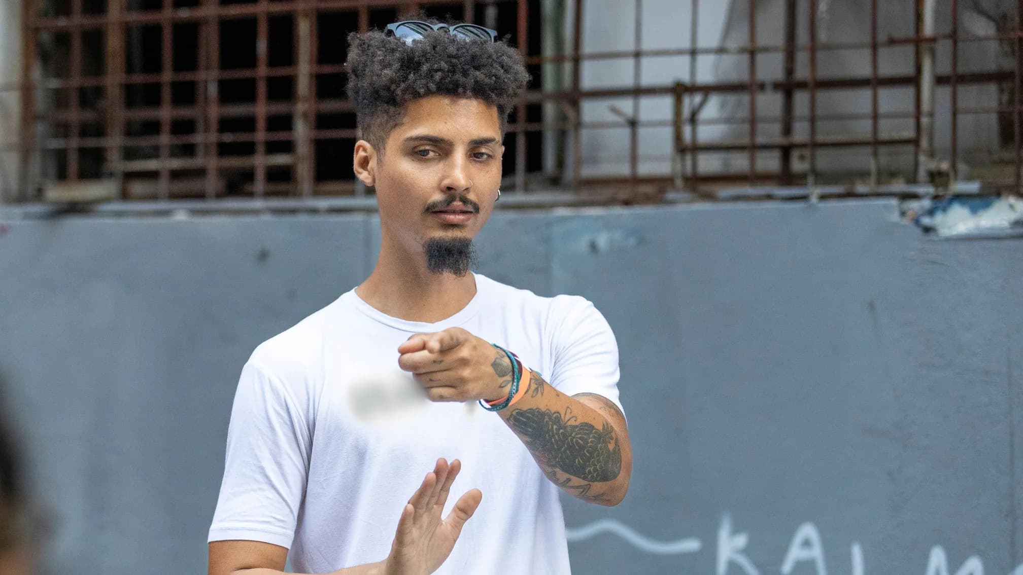 Jovem de camiseta branca sinaliza em Libras diante de um muro de rua, com um tambor ao lado